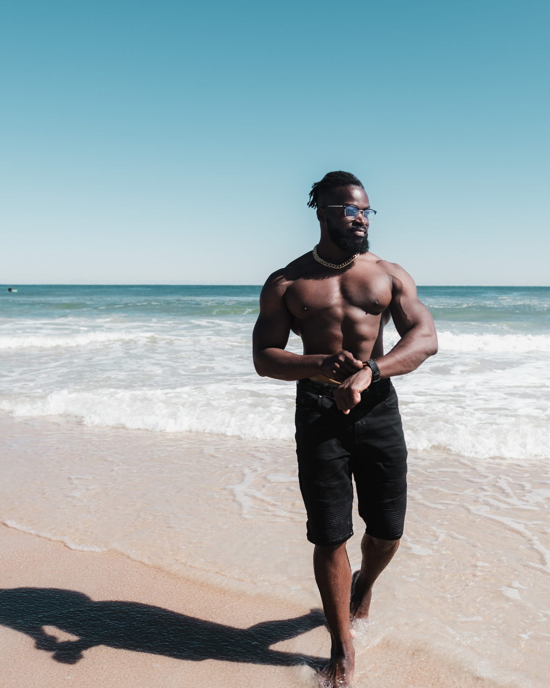 Shirtless, muscular man walking on the beach wearing black shorts and sunglasses